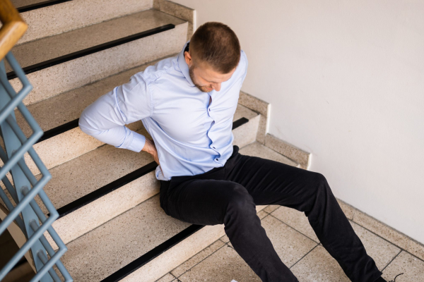 person in office attire rubbing their back while sitting at the bottom of a staircase